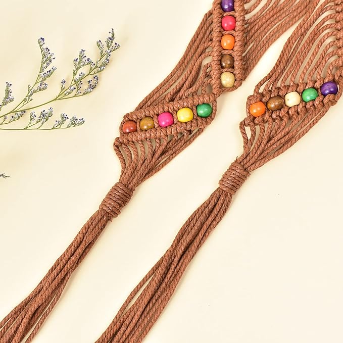 Ani Accessories Womens Belt Full view of a brown macrame tie belt with colorful wooden beads woven throughout, displayed on a light surface with dried decorative foliage to the left. The belt is tied in a prominent center knot with long tassels.
