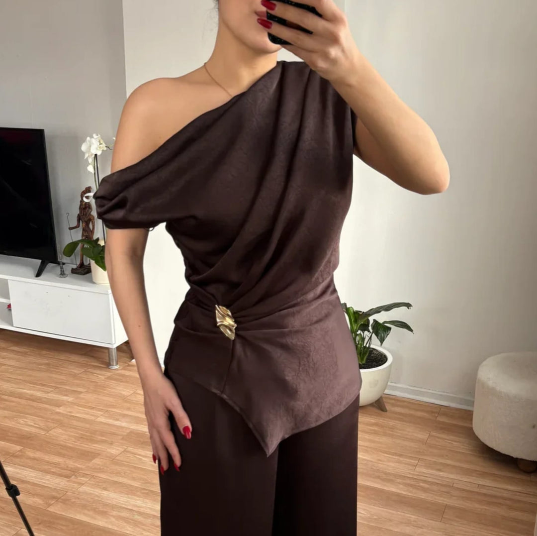 Woman wearing a gold abstract brooch on a brown one-shoulder draped top 