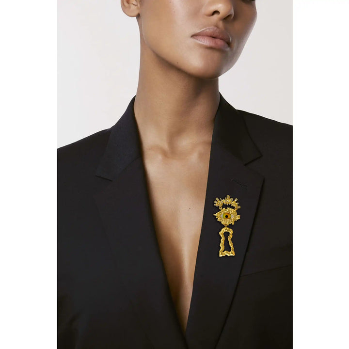 Woman wearing a gold surrealist eye brooch on a black blazer lapel with white color background 
