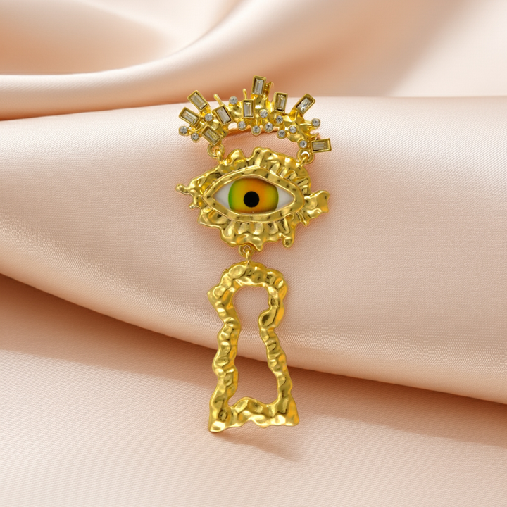 Gold surrealist eye brooch with green iris, spiky lashes and keyhole drop on pink satin 